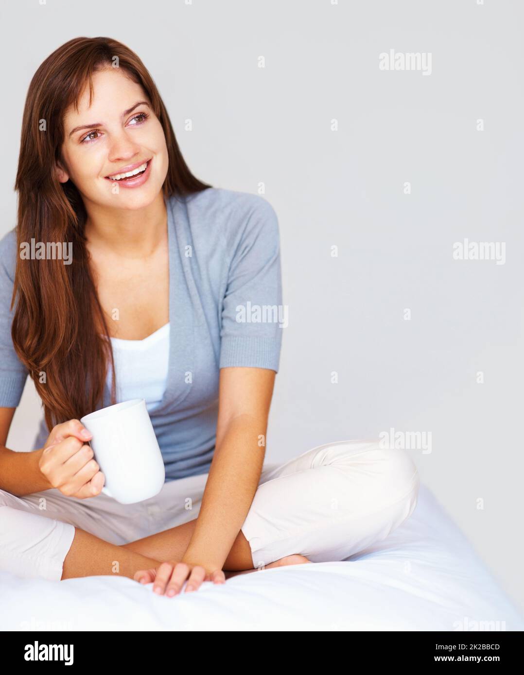 Nette, entspannte Frau, die Kaffee trinkt und in Gedanken wegschaut. Glückliche, nachdenkliche Frau, die die Beine gekreuzt auf dem Bett sitzt und eine Tasse Kaffee trinkt. Stockfoto