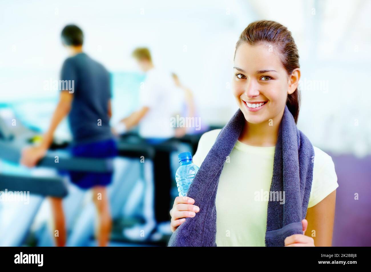 Junge Frau lächelt. Porträt einer jungen lächelnden Frau, die eine Wasserflasche und ein Handtuch hält, und Menschen, die im Hintergrund arbeiten. Stockfoto