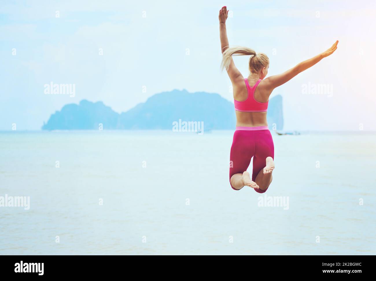 Fühlen Sie sich fit und fabelhaft. Rückansicht einer sportlichen jungen Frau, die an einem tropischen Strand in die Luft springt. Stockfoto