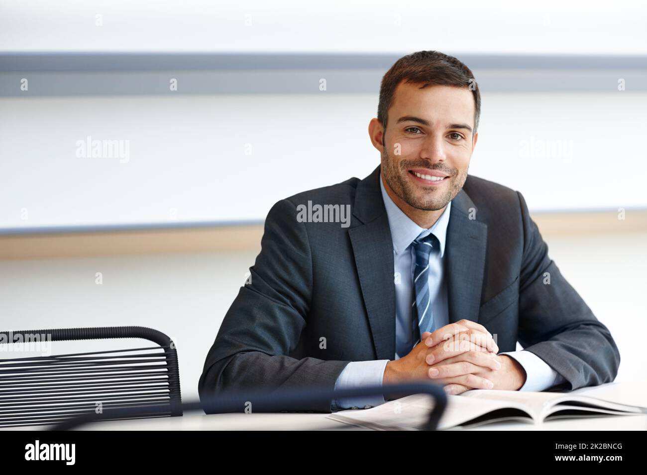 Bereit zu helfen. Ein hübscher junger Geschäftsmann, der mit einem Lächeln an seinem Schreibtisch sitzt. Stockfoto