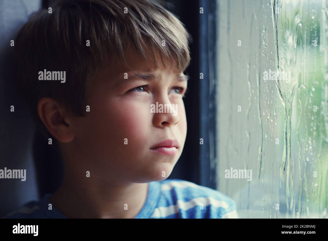 Gelangweilt, sich zu langweilen. Aufnahme eines traurigen Jungen, der den Regen durch ein Fenster zu Hause beobachtete. Stockfoto