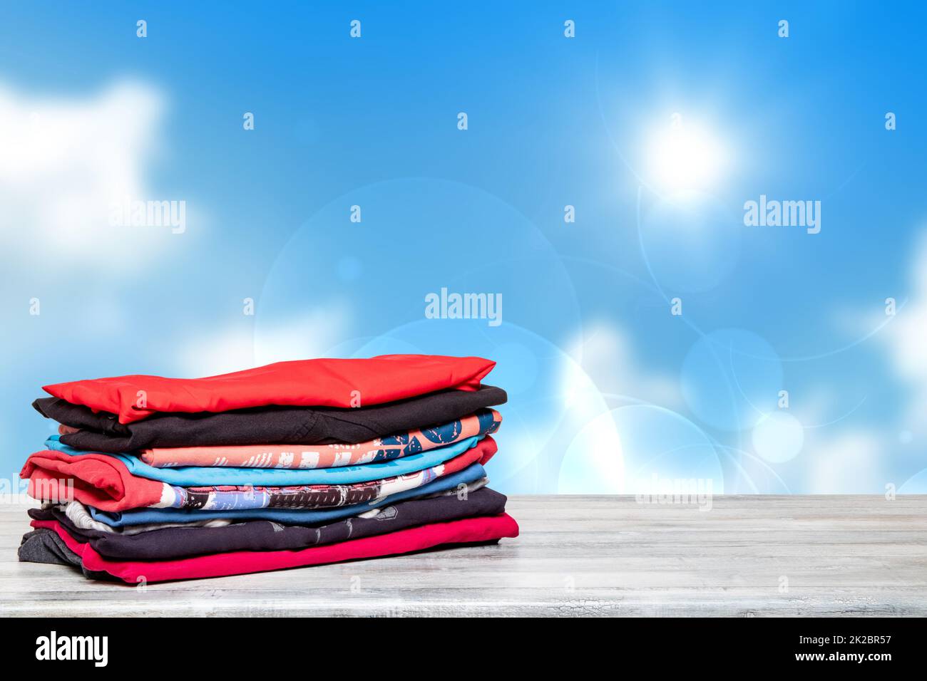 Gestaple farbenfrohe Kleidung. Nahaufnahme eines Stapels bunter T-Shirts oder Shirts auf einem hellen Tisch vor abstraktem, unscharfem sonnigen blauen Himmelshintergrund. Platz für die Montage Ihres Produktdisplays. Sommermode. Stockfoto