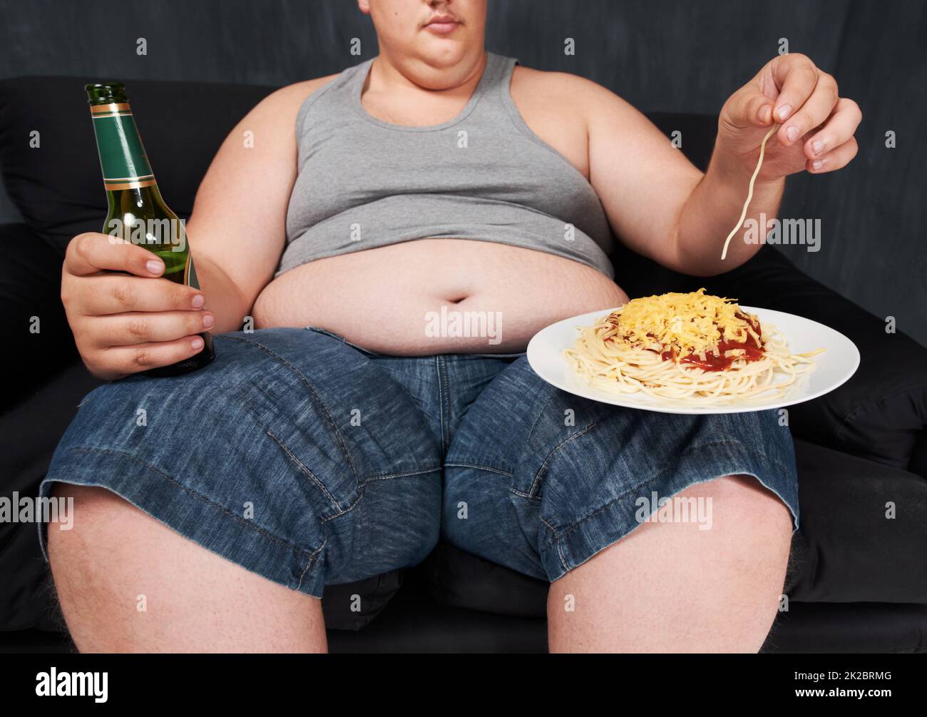 Wer braucht eine Gabel. Eine ausgeschnittene Aufnahme eines übergewichtigen jungen Mannes, der auf einem Sofa sitzt und Spaghetti mit den Händen isst. Stockfoto