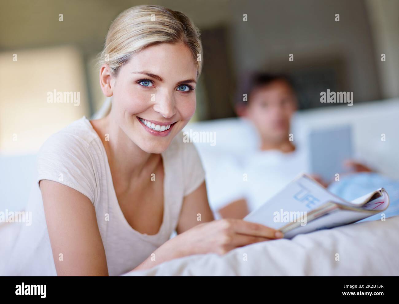 Blättern Sie um für ein glücklicheres Ich. Aufnahme einer glücklichen jungen Frau, die im Bett liegt und eine Zeitschrift liest, während ihr Mann ein digitales Tablet im Hintergrund verwendet. Stockfoto