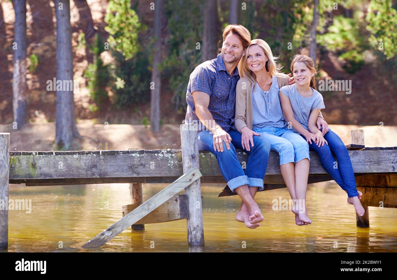 Die Zeit mit der Familie ist so kostbar. Eine glückliche dreiköpfige Familie sitzt zusammen auf dem Steg am See. Stockfoto