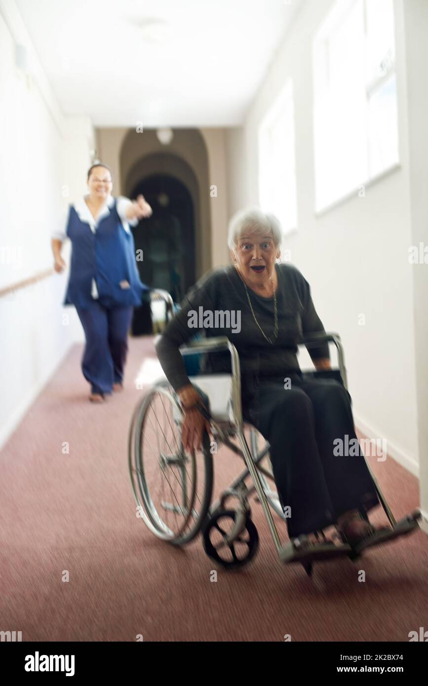 Ich bin aus diesem Gelenk herausgekommen. Aufnahme einer schelmischen älteren Frau, die ihren Rollstuhl benutzte, um in einem Altersheim von ihrer Krankenschwester wegzu laufen. Stockfoto