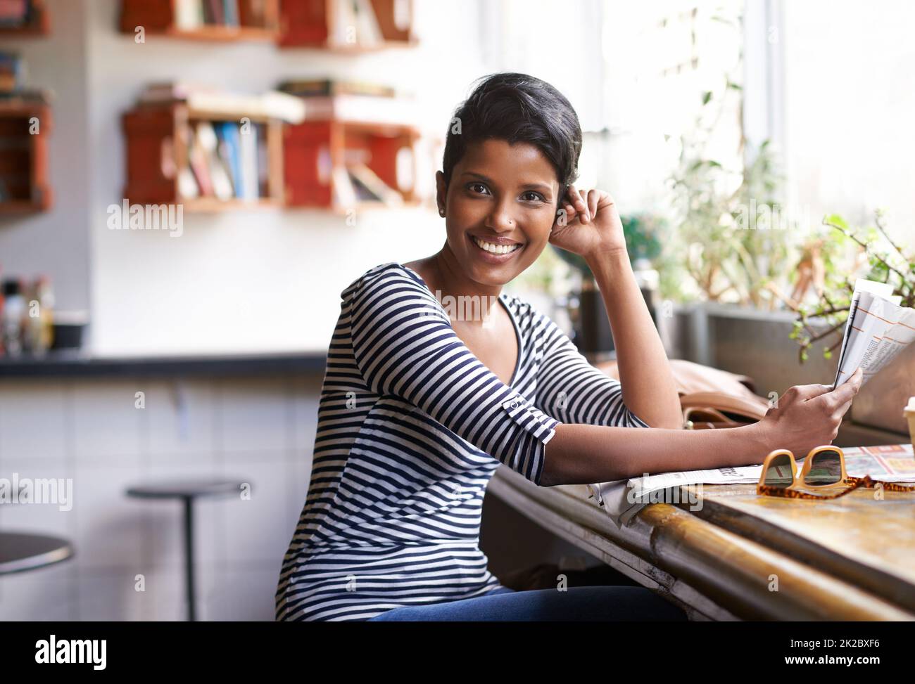 Mit ihrem Kaffee einige Neuigkeiten einzufangen. Eine junge Frau, die in einem Café mit einer Zeitung in der Hand sitzt. Stockfoto