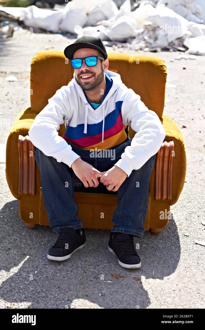 Hip und Happening. Ein trendiger Mann mit Sonnenbrille und Mütze auf lächelnd und sitzt auf einer Couch im Freien. Stockfoto