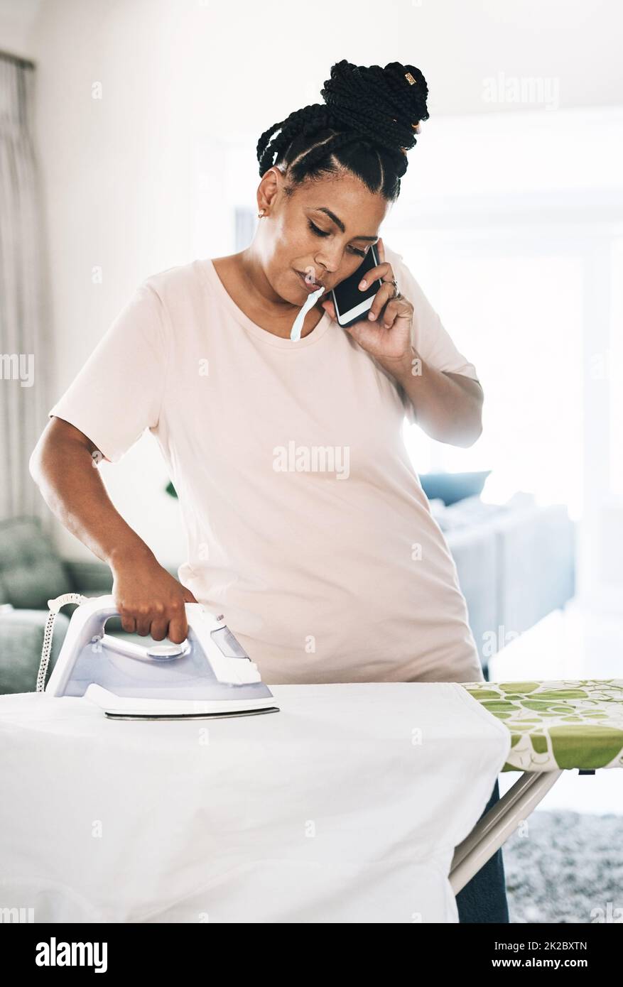 Multitasking ist eine Fähigkeit. Aufnahme einer jungen Frau, die zu Hause am Telefon Kleidung bügelt. Stockfoto