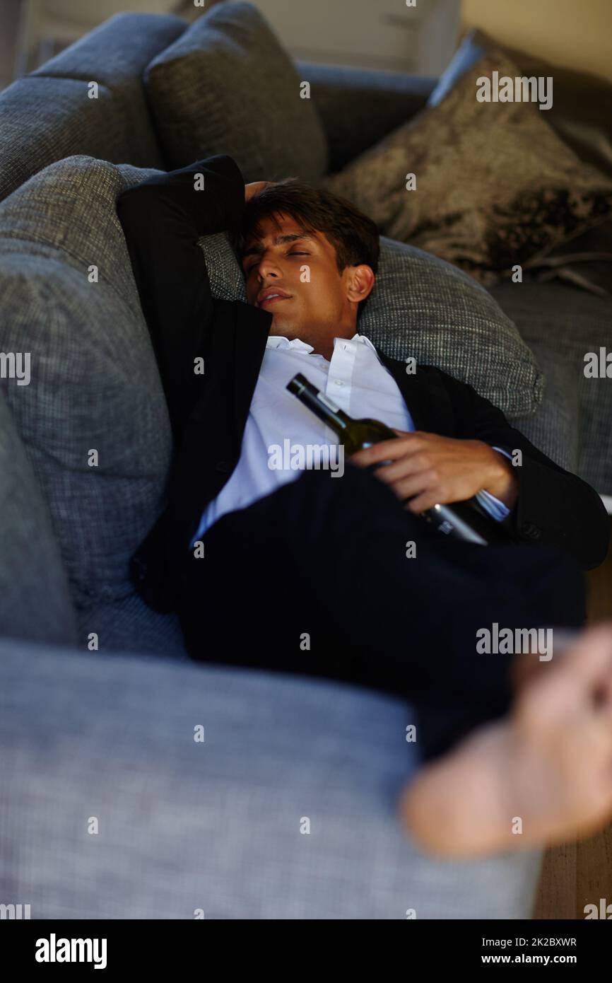 Der Kater schläft weg. Aufnahme eines jungen Mannes, der ohnmächtig auf einem Sofa lag, während er eine Flasche Wein in der Hand hielt. Stockfoto