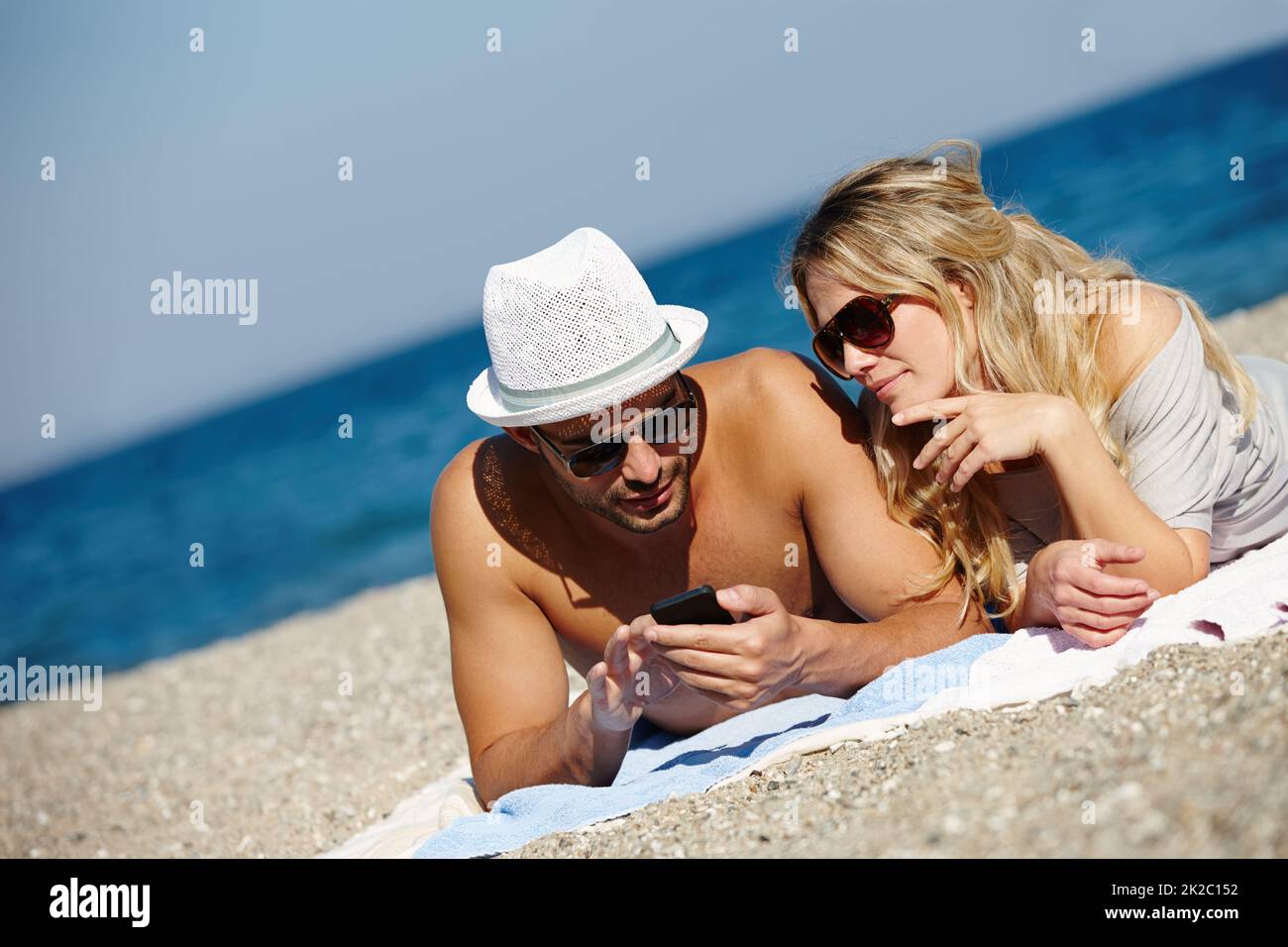 Nie war es einfacher, in Kontakt zu bleiben. Aufnahme eines jungen Paares, das ein Handy benutzte, während es sich am Strand sonnen hat. Stockfoto