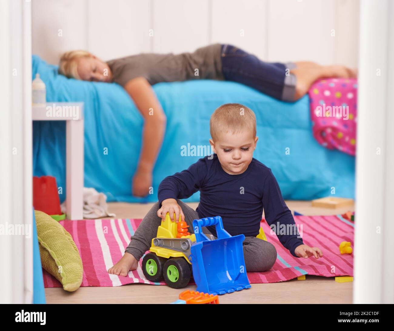 Elternschaft ist anstrengend. Ein Kleinkind spielt mit seinen Spielsachen, während seine Mutter auf seinem Bett nippt. Stockfoto