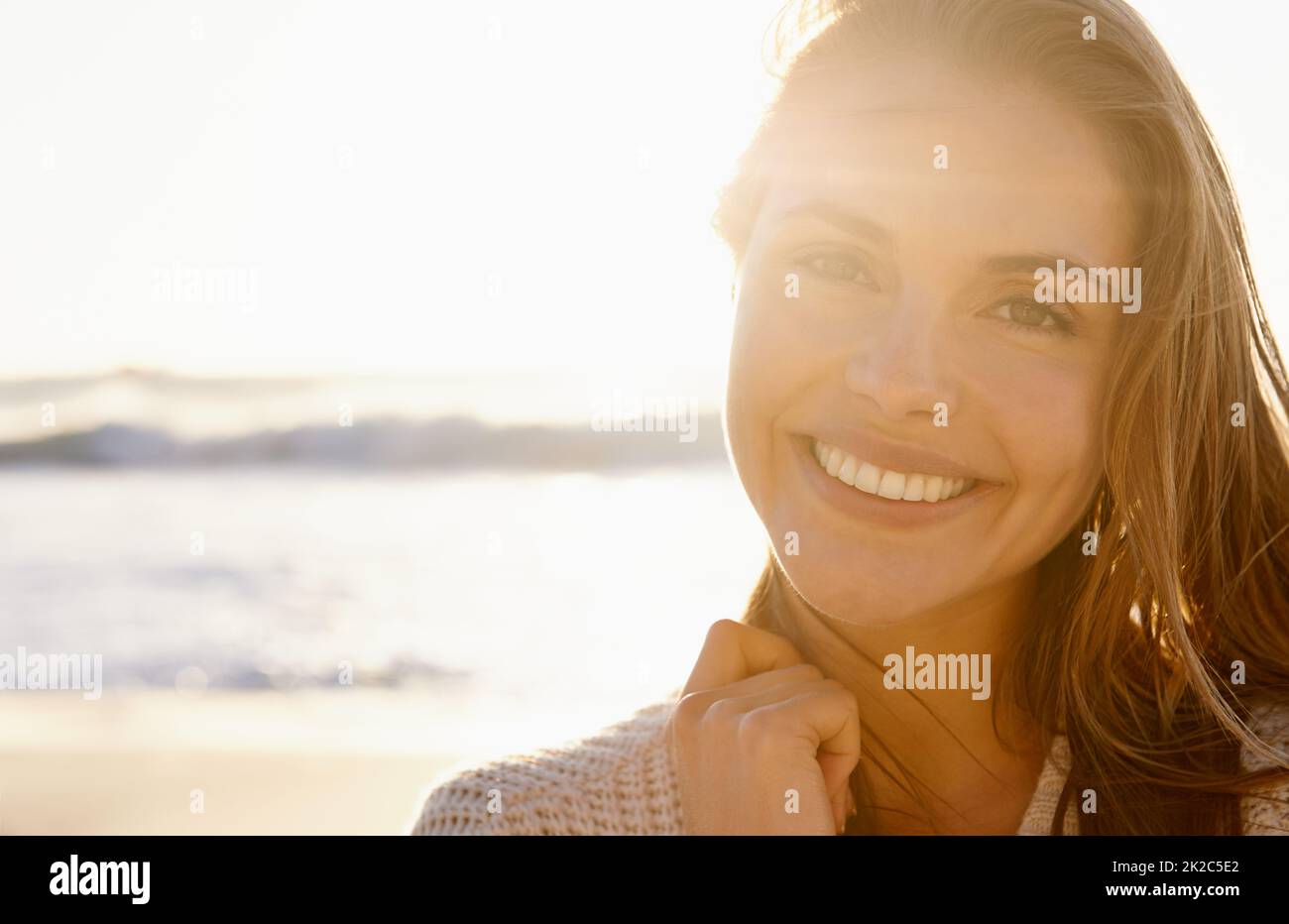 Atemberaubendes Lächeln bei Sonnenuntergang. Eine hübsche junge Frau, die selbstbewusst lächelt, als die Sonne im Hintergrund am Strand untergeht. Stockfoto