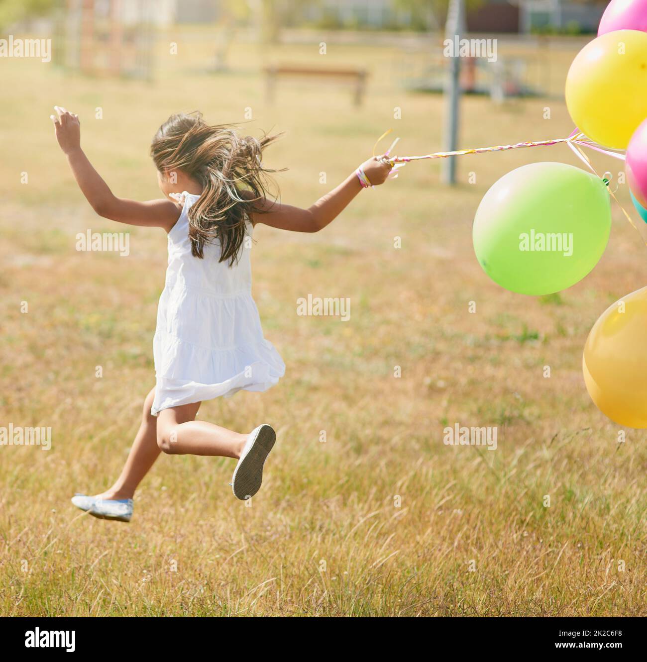 Glücklich hüpfend. Rückansicht eines kleinen Mädchens, das mit einer Reihe von Luftballons draußen durch ein Feld läuft. Stockfoto