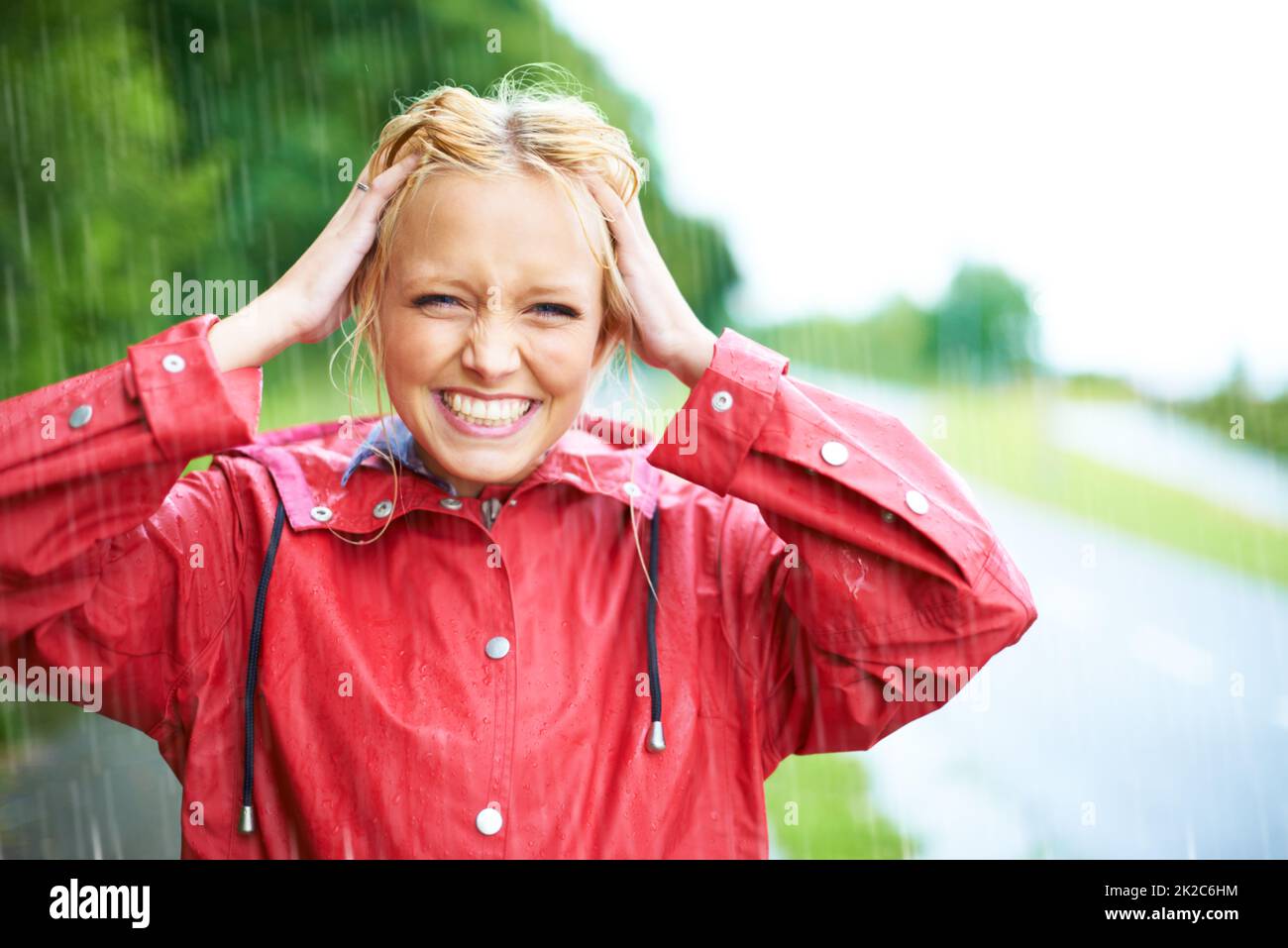 Nichts ist besser als ein wenig frisches Regenwasser. Wunderschöne junge blonde Frau trägt einen roten Regenmantel im Regen draußen auf einer Landstraße. Stockfoto