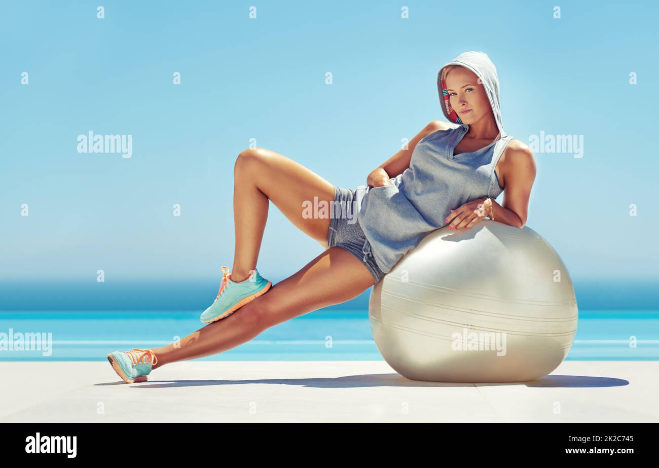 Sich der Fitness verschrieben. Porträt einer schönen jungen Frau, die sich am Pool auf einen Übungsball stützt. Stockfoto