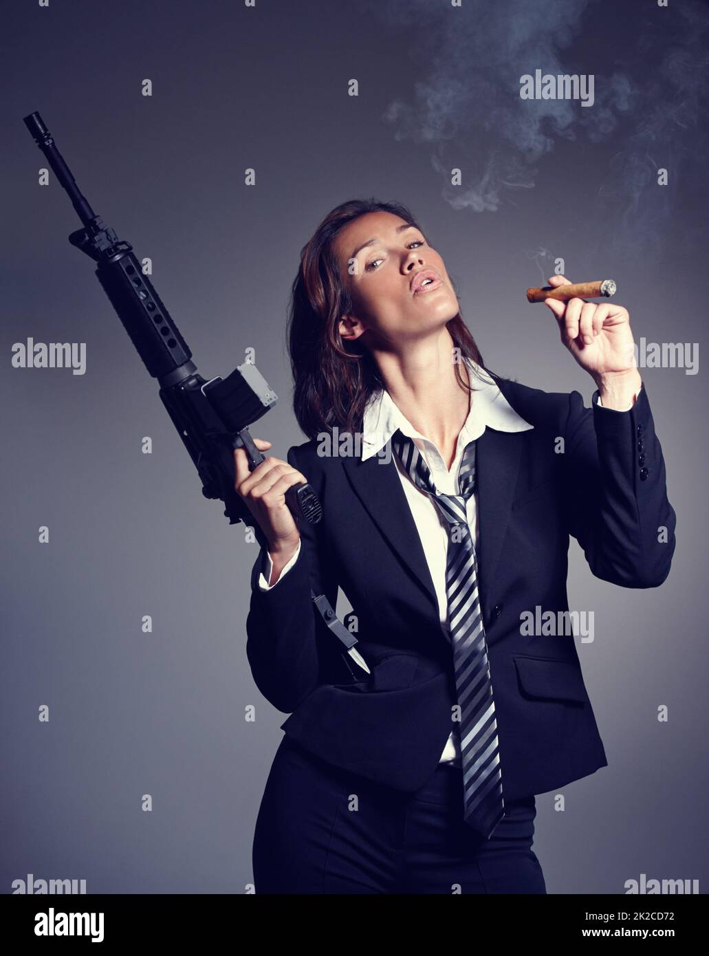 In Rauch aufgehen. Eine junge Geschäftsfrau in Anzug und Krawatte, die ein Gewehr hält, während sie eine Zigarre raucht. Stockfoto