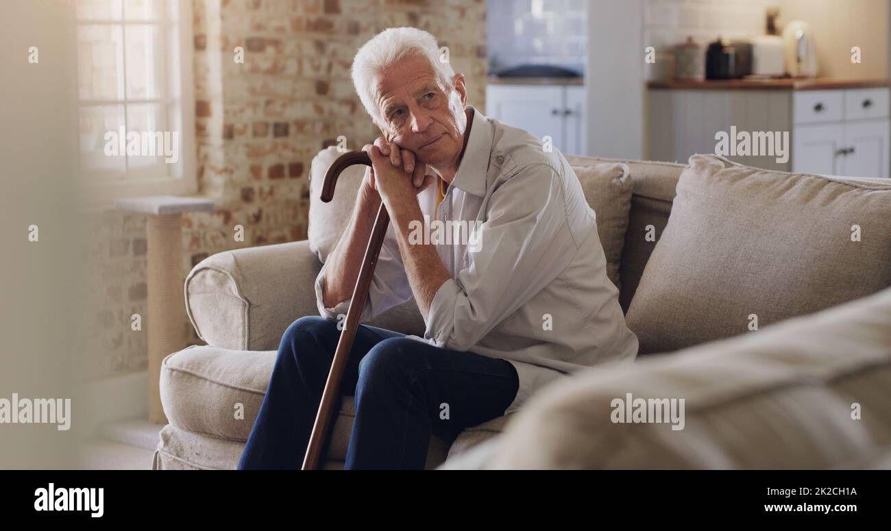 Wo war die ganze Zeit hin? Aufnahme eines älteren Mannes, der allein auf dem Sofa zu Hause sitzt und besinnlich aussieht, während er seinen Spazierstock hält. Stockfoto
