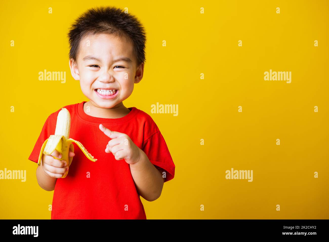 Kind niedlichen kleinen Jungen attraktives Lächeln trägt rote T-Shirt spielen Hält geschälte Banane zum Essen Stockfoto