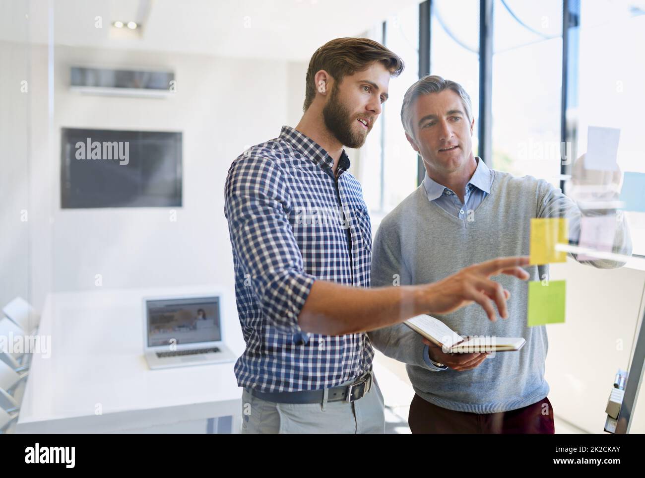 Sie glauben an ständige Innovation, um ihre Kunden zu beeindrucken. Aufnahme von zwei männlichen Kollegen, die während eines Brainstormings Haftnotizen an einer Glaswand anordnen. Stockfoto