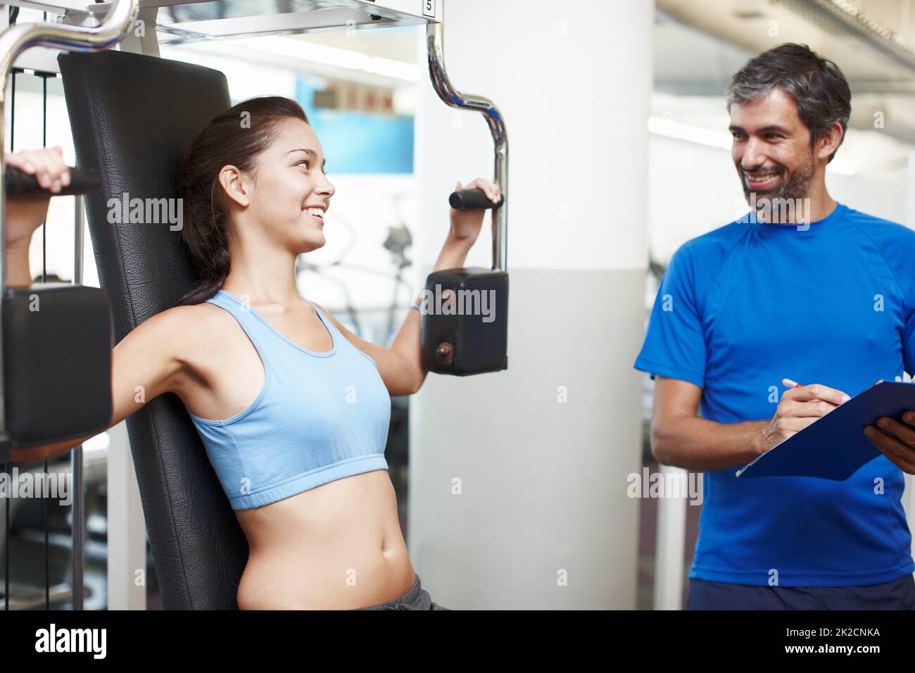 Mache ich das richtig? Eine kurze Aufnahme einer attraktiven jungen Frau, die mit einem Trainingsgerät unterwegs ist, während ihr persönlicher Trainer anschaut. Stockfoto