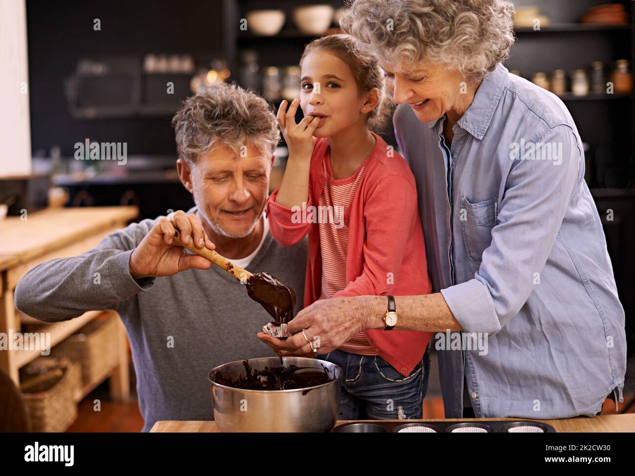 MMM... das ist so gut. Porträt eines kleinen Mädchens und ihrer Großeltern, die in der Küche Cupcakes machen. Stockfoto