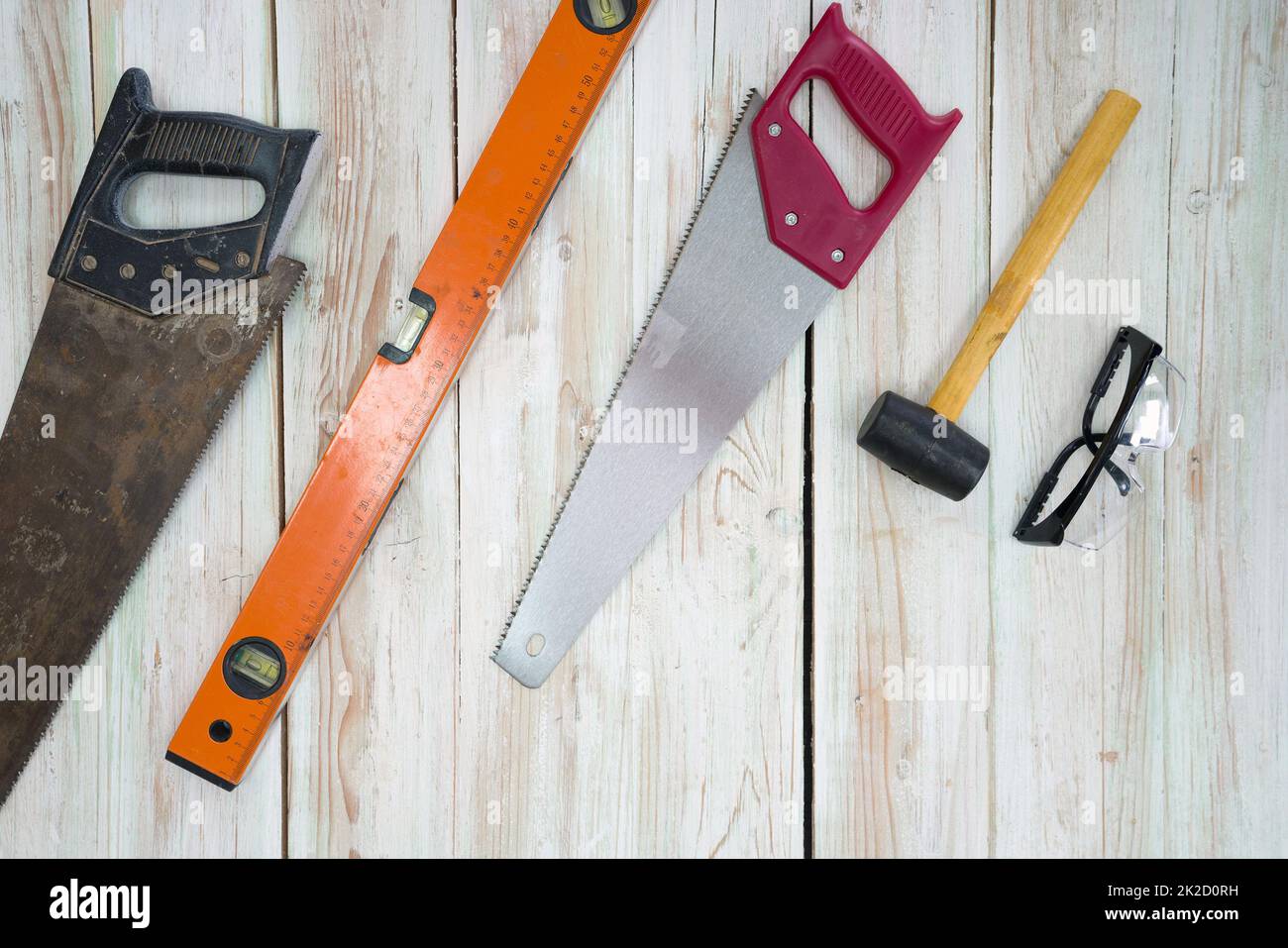 Die Ausrüstung für die Holzbearbeitung ist übersichtlich auf dem Holzboden angeordnet. Bestellung wie folgt: Alte Säge voller Rost, orangefarbene magnetische Aluminiumwaage, rote Handsäge, gelber Hammer, schwarze staubdichte Brille. Stockfoto