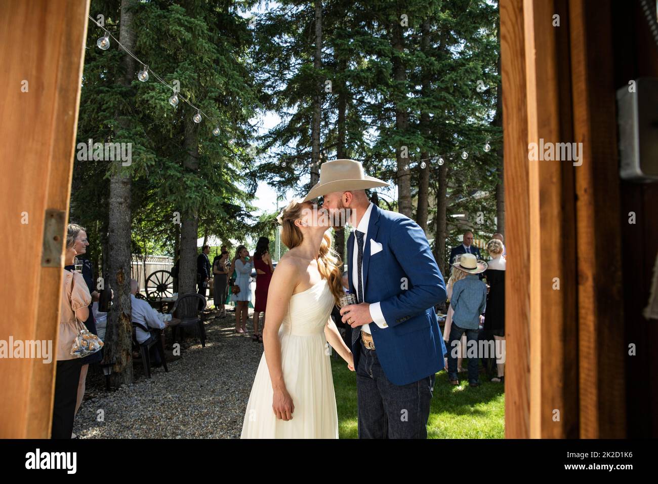 Braut und Bräutigam im Cowboy-Hut küssen sich am Hochzeitstag im sonnigen Garten Stockfoto