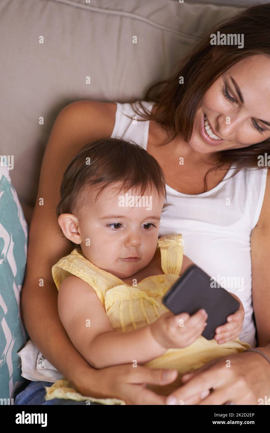 Es ist so neugierig. Ein Mädchen, das auf dem Schoß ihrer Mutter liegt und mit einem Handy spielt. Stockfoto