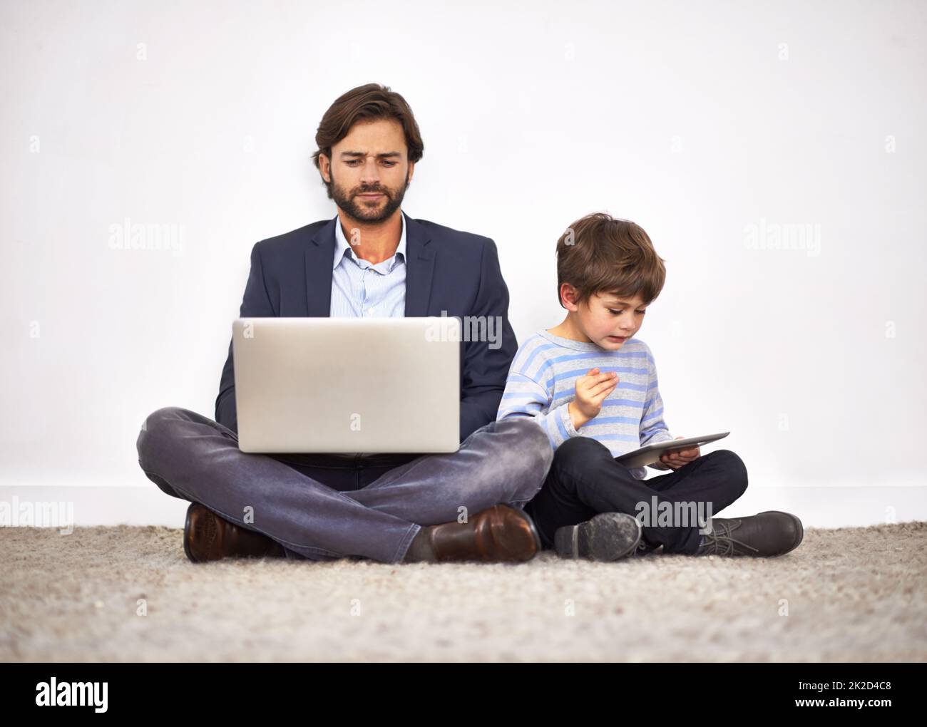 Jungen lieben ihre digitalen Spielzeuge. Ein Vater und ein Sohn sitzen auf dem Boden gegen eine Wand mit einem Laptop und einem digitalen Tablet. Stockfoto