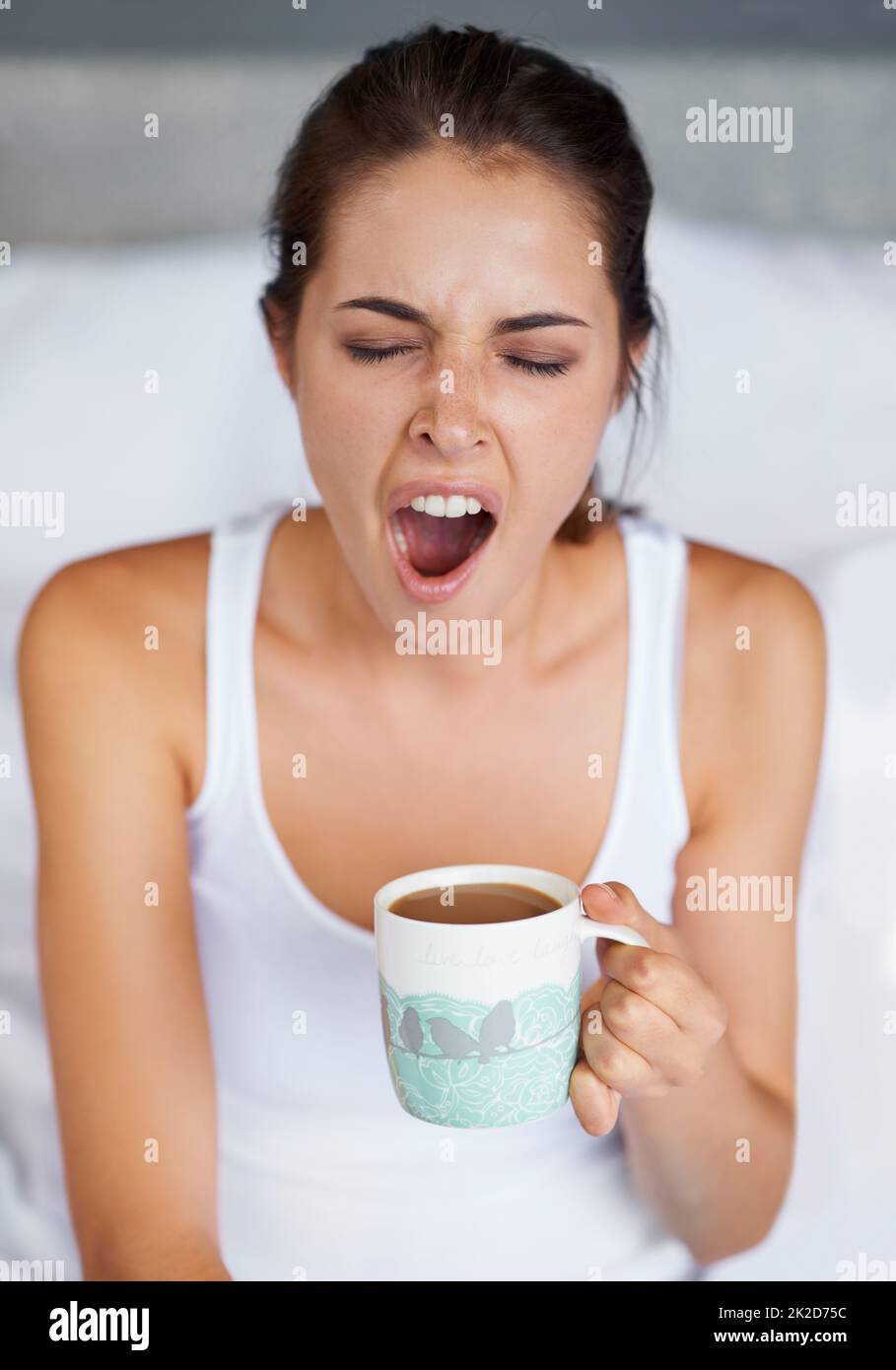 Ein großes GÄHNEN. Eine junge Frau, die ein großes Gähnen gibt, während sie mit einer Tasse Kaffee im Bett sitzt. Stockfoto