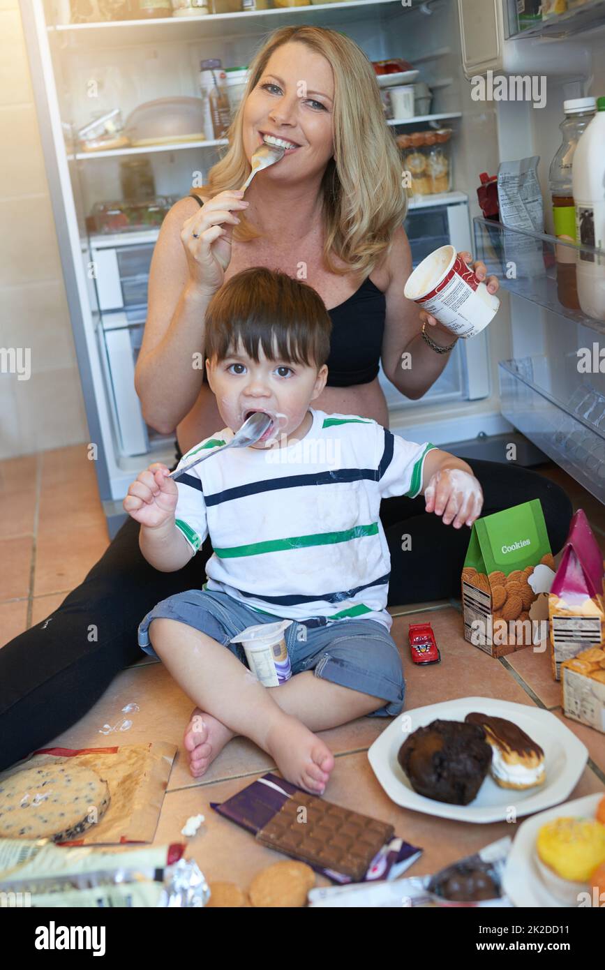 Manchmal wird das Verlangen besser von Ihnen. Porträt einer Schwangeren und ihres Babys, die Leckereien essen, während sie auf dem Küchenboden sitzen. Stockfoto