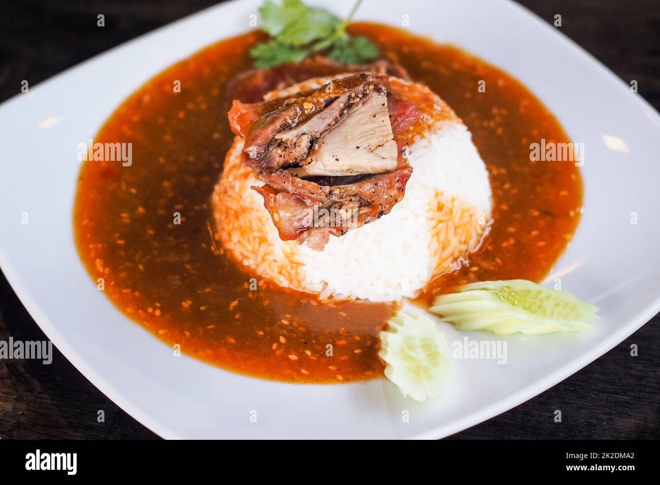 Rotes gegrilltes Schweinefleisch auf Reis, Speisen mit süßem Curry und Gemüse (Gurke, Koriander) auf weißer Keramikplatte servieren. Stockfoto