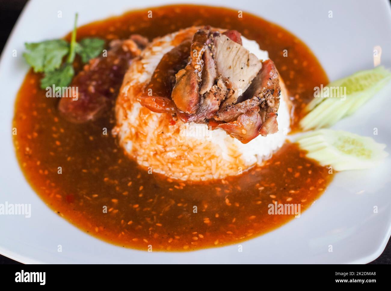 Rotes gegrilltes Schweinefleisch auf Reis, Speisen mit süßem Curry und Gemüse (Gurke, Koriander) auf weißer Keramikplatte servieren. Stockfoto