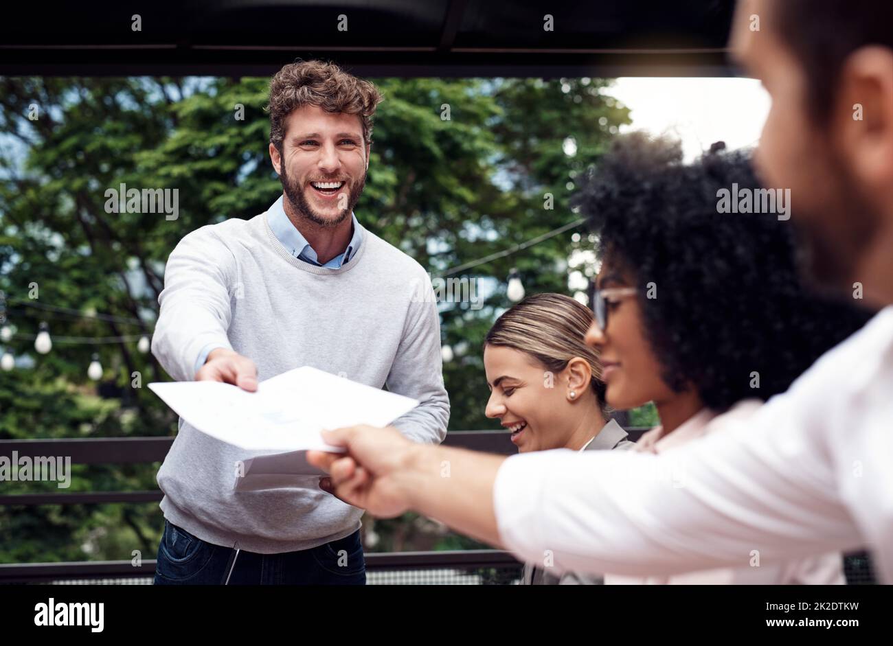 Bitte nehmen Sie eine und geben Sie sie weiter. Freigestellte Aufnahme eines gutaussehenden jungen Geschäftsmanns, der während eines Meetings im Freien steht und Dokumente an seine sitzenden Kollegen verteilt. Stockfoto