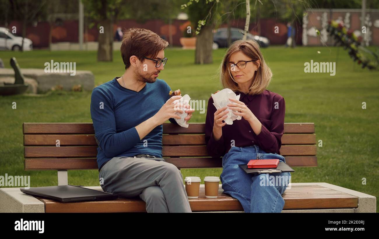 Hungriger junger Geschäftsmann genießt einen Snack. Essen Sie im Freien. Freunde, die einen Snack in einem Park zu sich nehmen. Freunde essen mit Gusto. Stockfoto