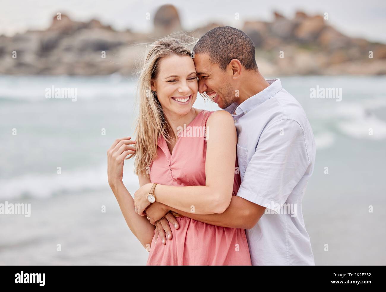 Reisen, Strand und Paar umarmen sich, glücklich und lachend, während sie sich im Freien verbinden und den Blick auf das Meer genießen. Liebe, Beziehung und Wasserspaß mit Entspannung Stockfoto