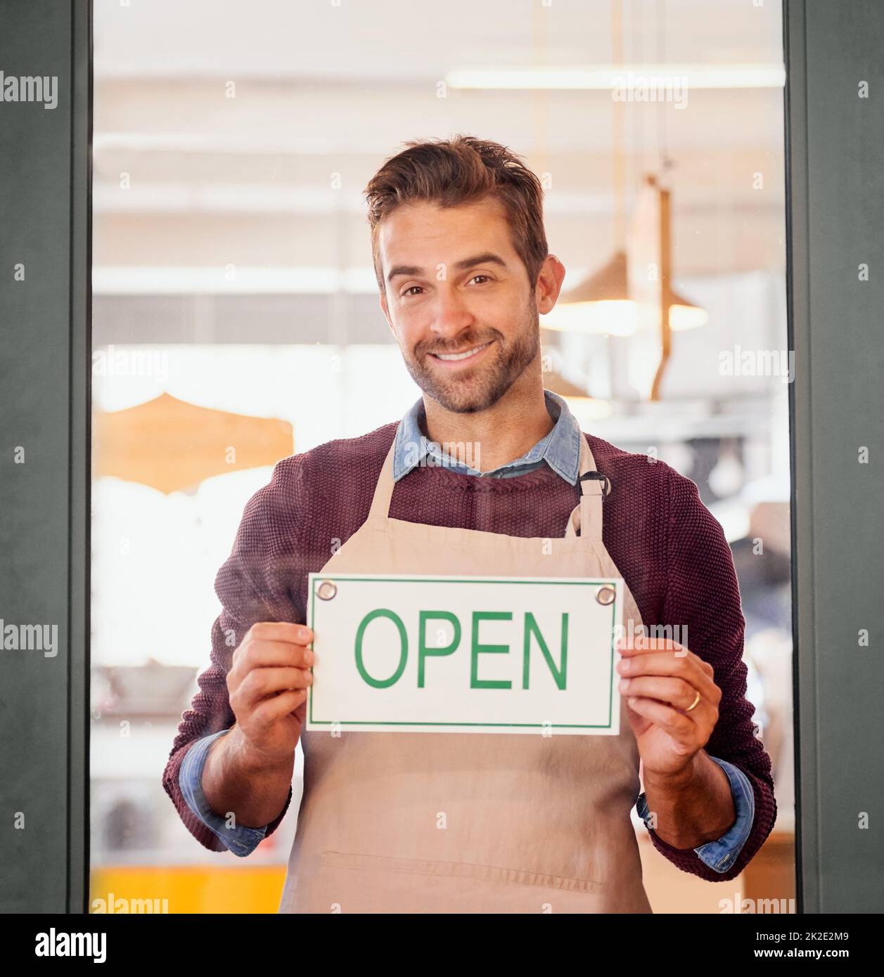Kommen Sie herein. Porträt eines jungen Geschäftsbesitzers, der mit einem offenen Schild in der Tür seines Cafés steht. Stockfoto