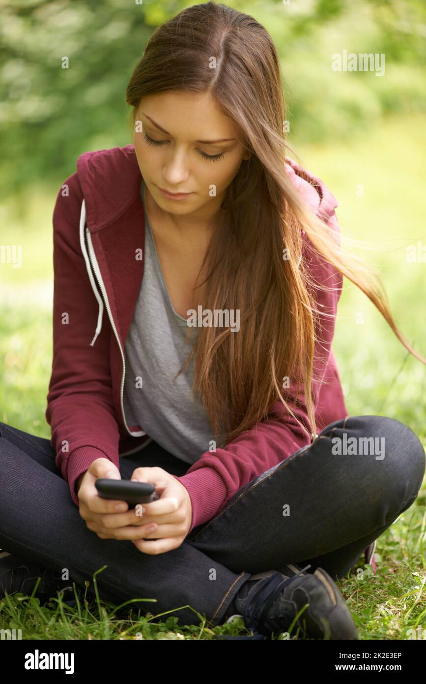 Immer verbunden. Ein hübsches junges Mädchen, das auf einem grünen Feld sitzt und eine SMS an ihr Telefon schreibt. Stockfoto