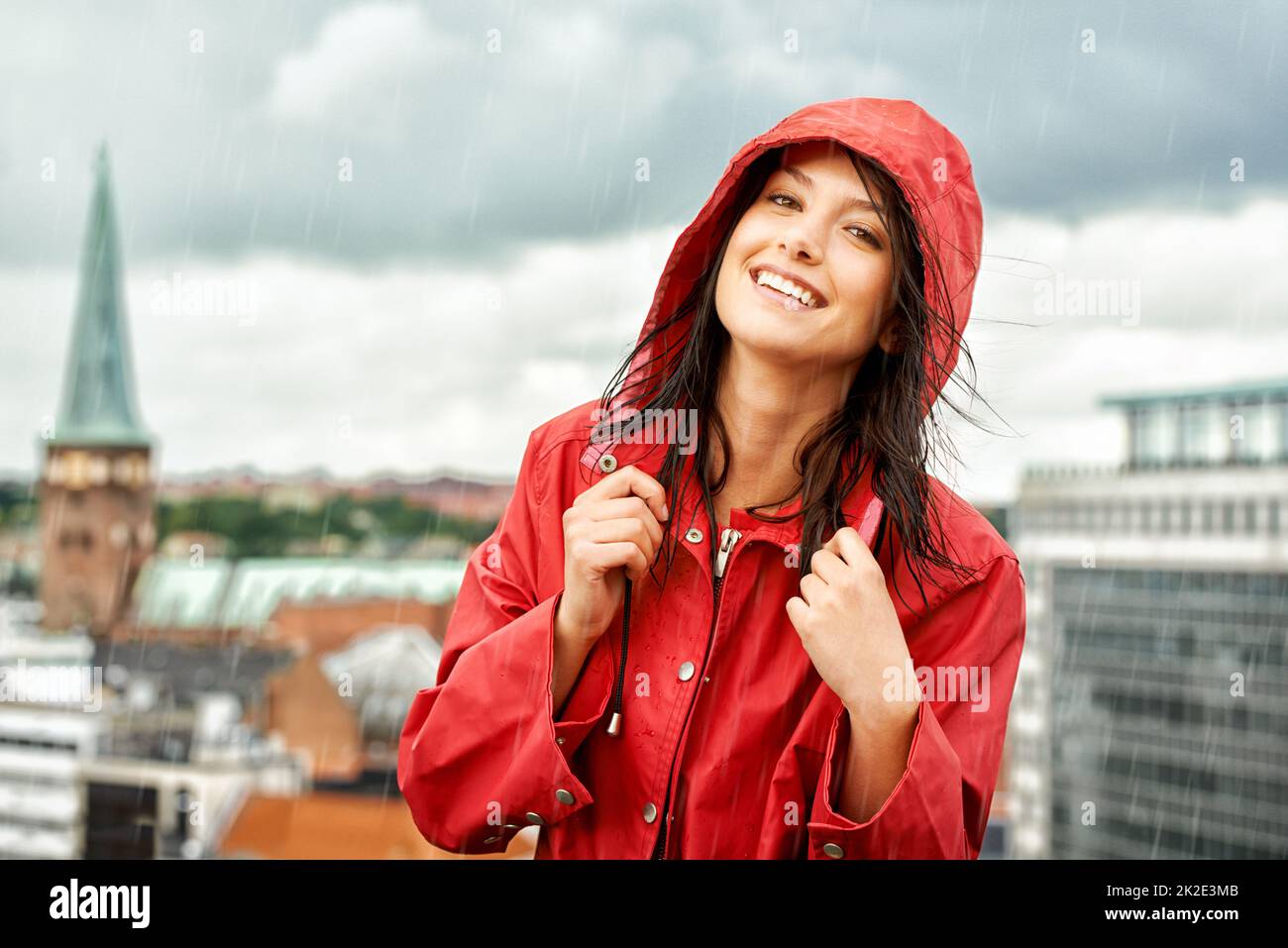 Ein regnerischer Tag kann sie nicht zum Boden bringen. Hübsche junge Frau, die lächelt, während sie einen roten Regenmantel trägt - Porträt. Stockfoto