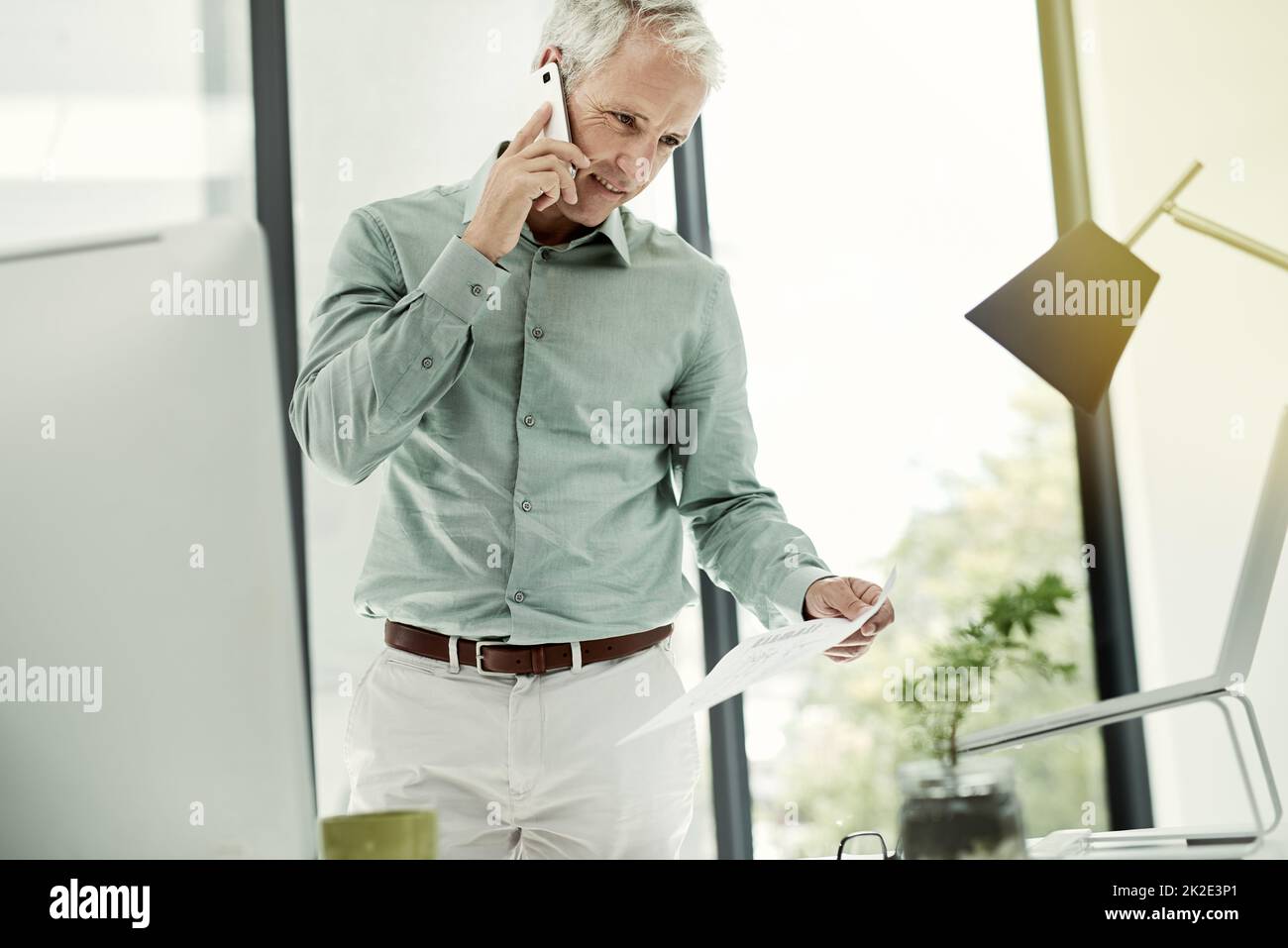 Im Gespräch über den Vertrag. Aufnahme eines reifen Geschäftsmannes, der am Telefon telefoniert, während er an seinem Schreibtisch in einem Büro steht. Stockfoto