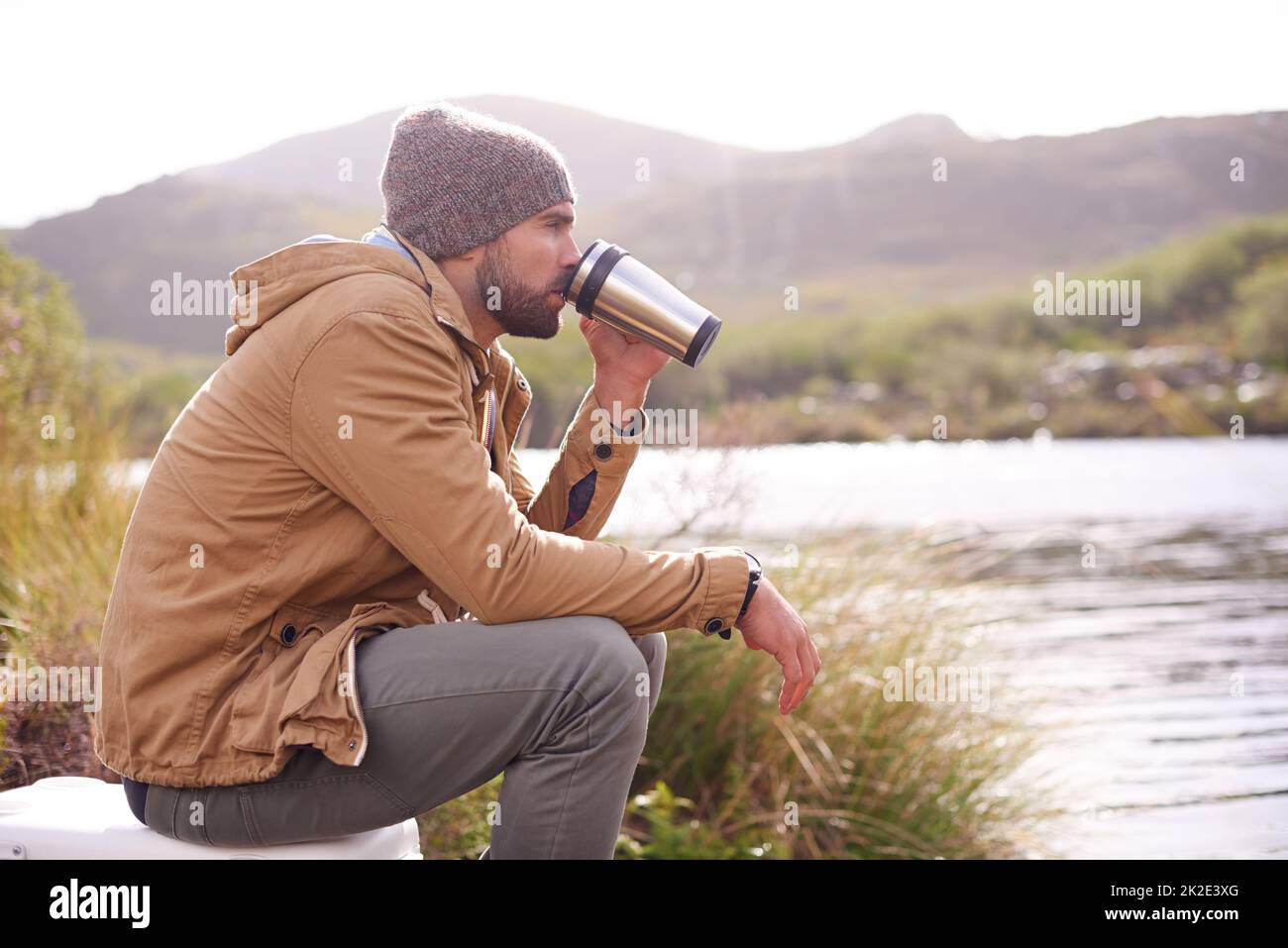 Genießen Sie die Landschaft. Aufnahme eines hübschen Mannes, der an einem See sitzt und aus einer Thermoskanne trinkt. Stockfoto