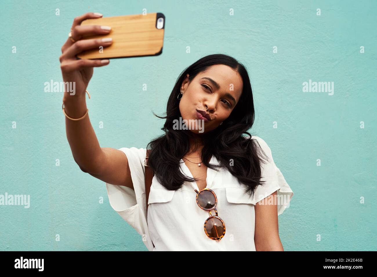 Wo wären Millennials ohne das Selfie? Abgeschnittene Aufnahme einer attraktiven jungen Frau, die mit ihrem Handy ein Selfie vor blauem Hintergrund macht. Stockfoto