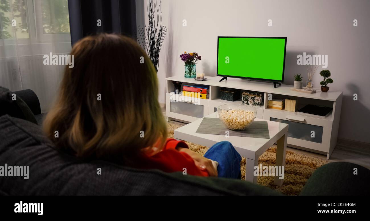 Ein Mädchen, das sich Green Chroma Key TV ansieht, entspannt auf einer Couch zu Hause sitzt. Frau im Wohnzimmer, die Sportwettkämpfe, Nachrichten, Sitcom TV-Sendungen oder einen Film auf einem grünen Bildschirm ansieht. Stockfoto