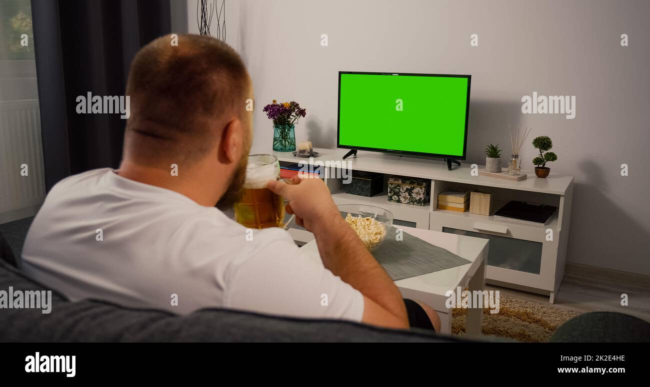 Mann, der zu Hause einen Fernseher mit grünem Bildschirm sieht. Trinken Sie Bier Essen Popcorn. Über der Schulter Rückansicht. Chroma-Taste. Stockfoto