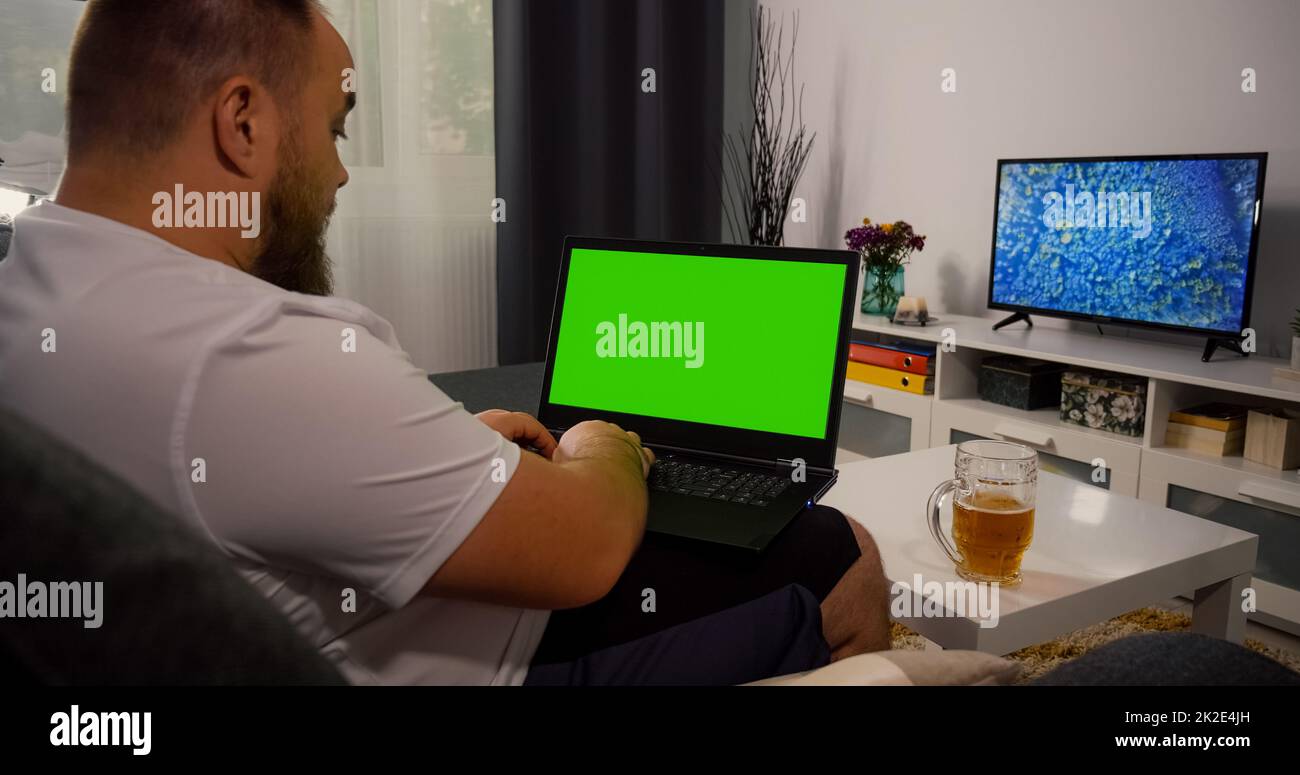 Mann, der auf dem Sofa sitzt und ein Notebook mit Greenscreen und Chroma Key benutzt. Modell mit Tracking Markers und Alpha Matte. Kaukasischer Männchen, der sich abends auf dem Laptop entspannt und Bier trinkt. Online-Suche. Stockfoto