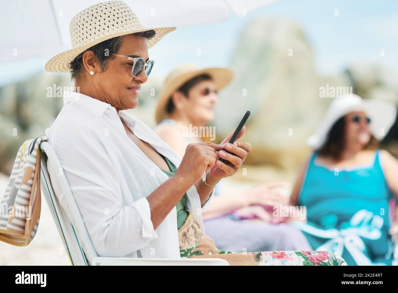 Schicken Sie all unsere Selfies an unsere Kinder. Aufnahme einer reifen Frau, die alleine sitzt und ihr Handy während eines Tages am Strand mit Freunden benutzt. Stockfoto