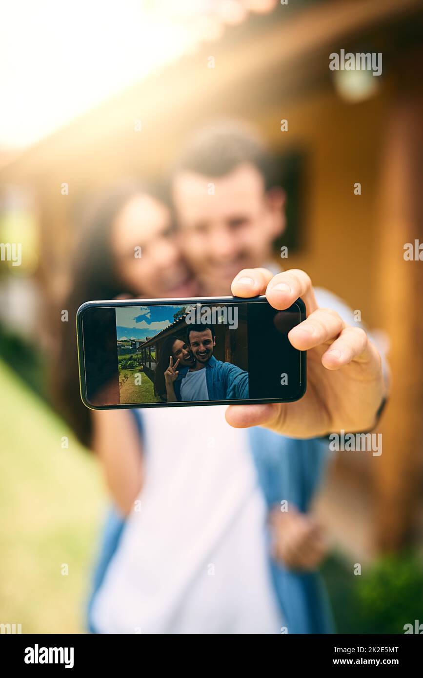 Ihre Liebe festhalten. Ausgeschnittene Aufnahme eines liebevollen jungen Paares, das draußen Selfies mit seinem Haus im Hintergrund macht. Stockfoto