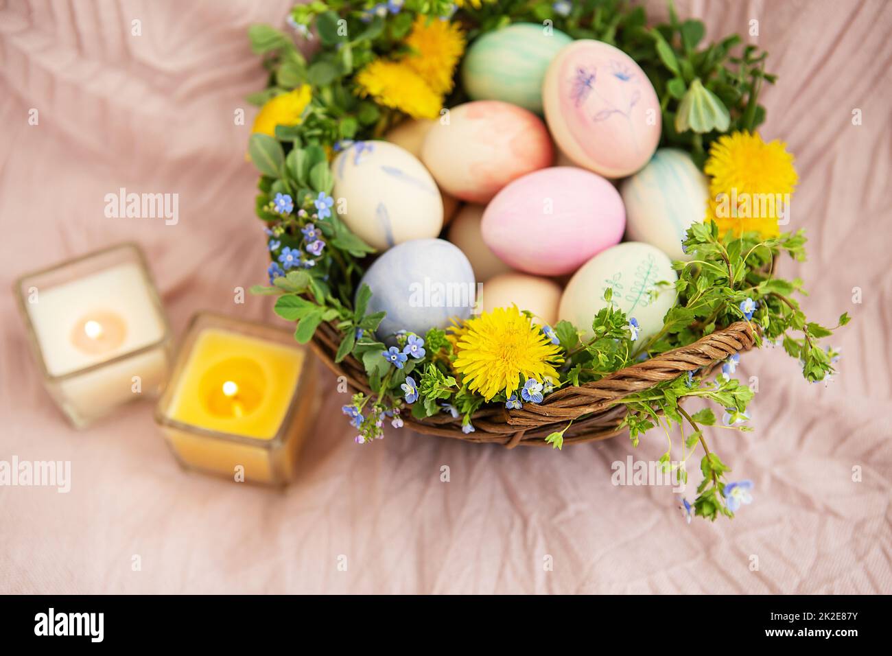 Auf einer pastellfarbenen Bettdecke befindet sich ein Korb mit Gras und leuchtenden Blumen. Der Korb enthält Ostereier, die mit Aquarellen in wunderschönen Pastelltönen bemalt sind. Stockfoto