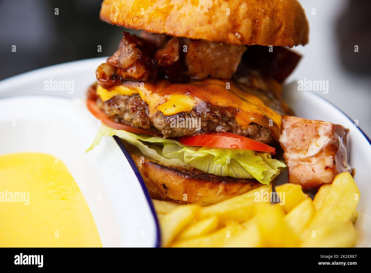 Das Konzept von Fast Food und Essen zum Mitnehmen. Ein saftiger Hamburger liegt neben pommes frites auf einem Metallteller zusammen mit Cheddar Cheese Sauce im Hintergrund. Stockfoto